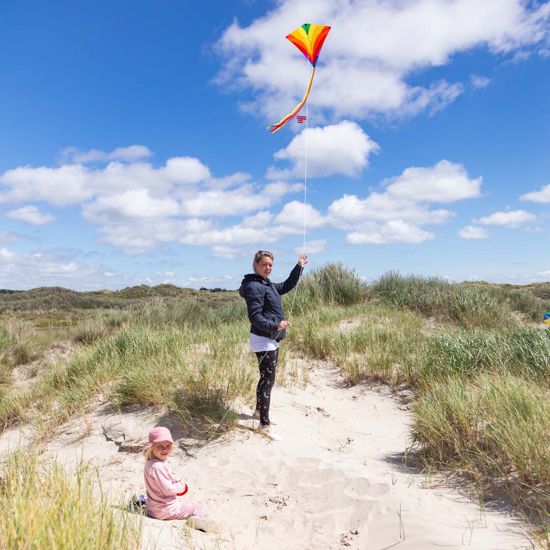 Vuxen och barn som flyger en färgglad Eddy Rainbow drake med lång gul svans och vitt snöre bland sanddyner vid stranden
