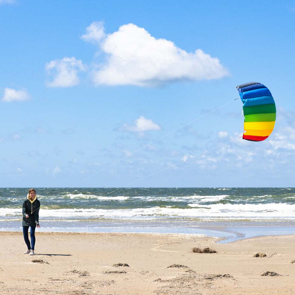 En ung kvinna står på en sandstrand vid havet och flyger en stor regnbågsfärgad Symphony-drake mot en blå himmel med vita moln.