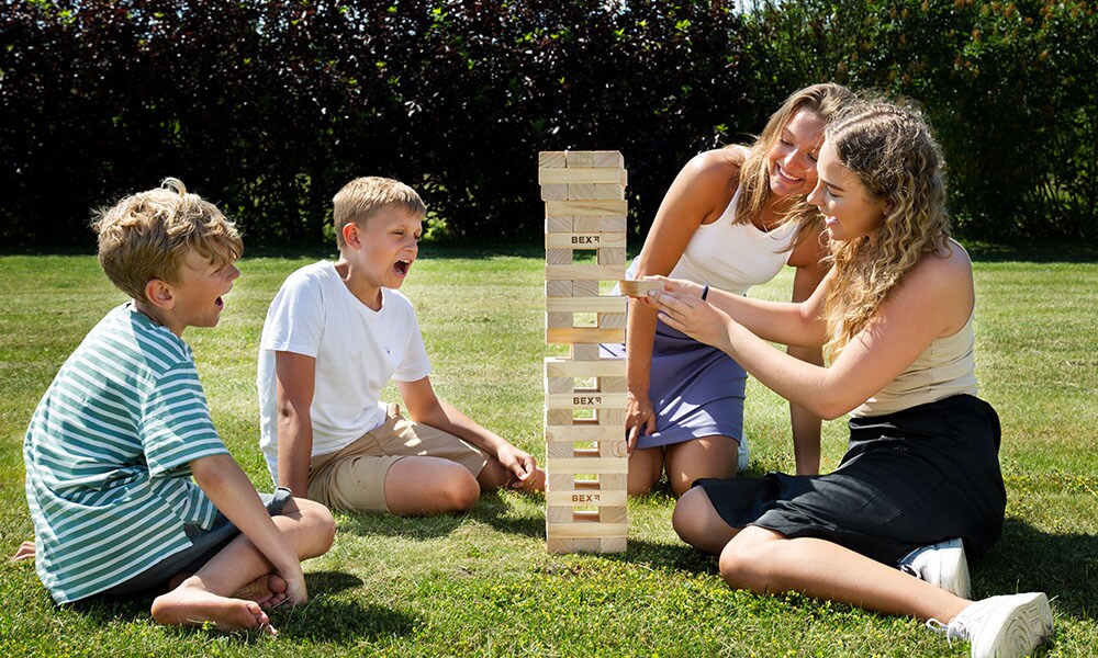 Två pojkar och två flickor spelar Giant Tower från BEX utomhus på en gräsmatta. En av pojkarna gapar av spänning medan en flicka drar ut en kloss