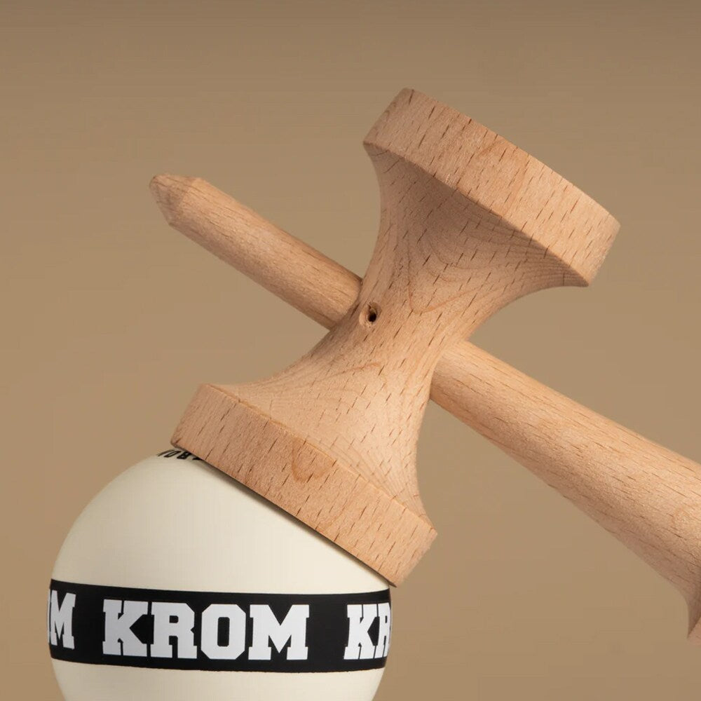 Närbild på en KROM Strogo Neapolitan kendama där den vita bollen (tama) med det svarta ”KROM”-bandet balanseras mot kanten av en av träkopparna på den ljusa ken-delen mot en beige bakgrund.