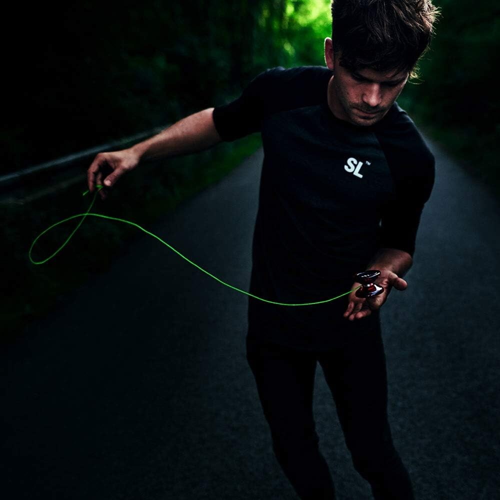 En ung man i mörk t-shirt utför ett yoyotrick med en röd och svart yoyo och ett neongrönt snöre på en mörk asfalterad väg.