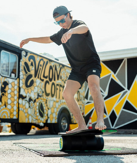 Man balanserar barfota på en Rockerboard med roller på en matta, med en färgglad food truck i bakgrunden