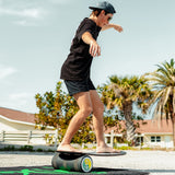 Person i shorts och keps balanserar på en Indoboard med roller på en matta utomhus