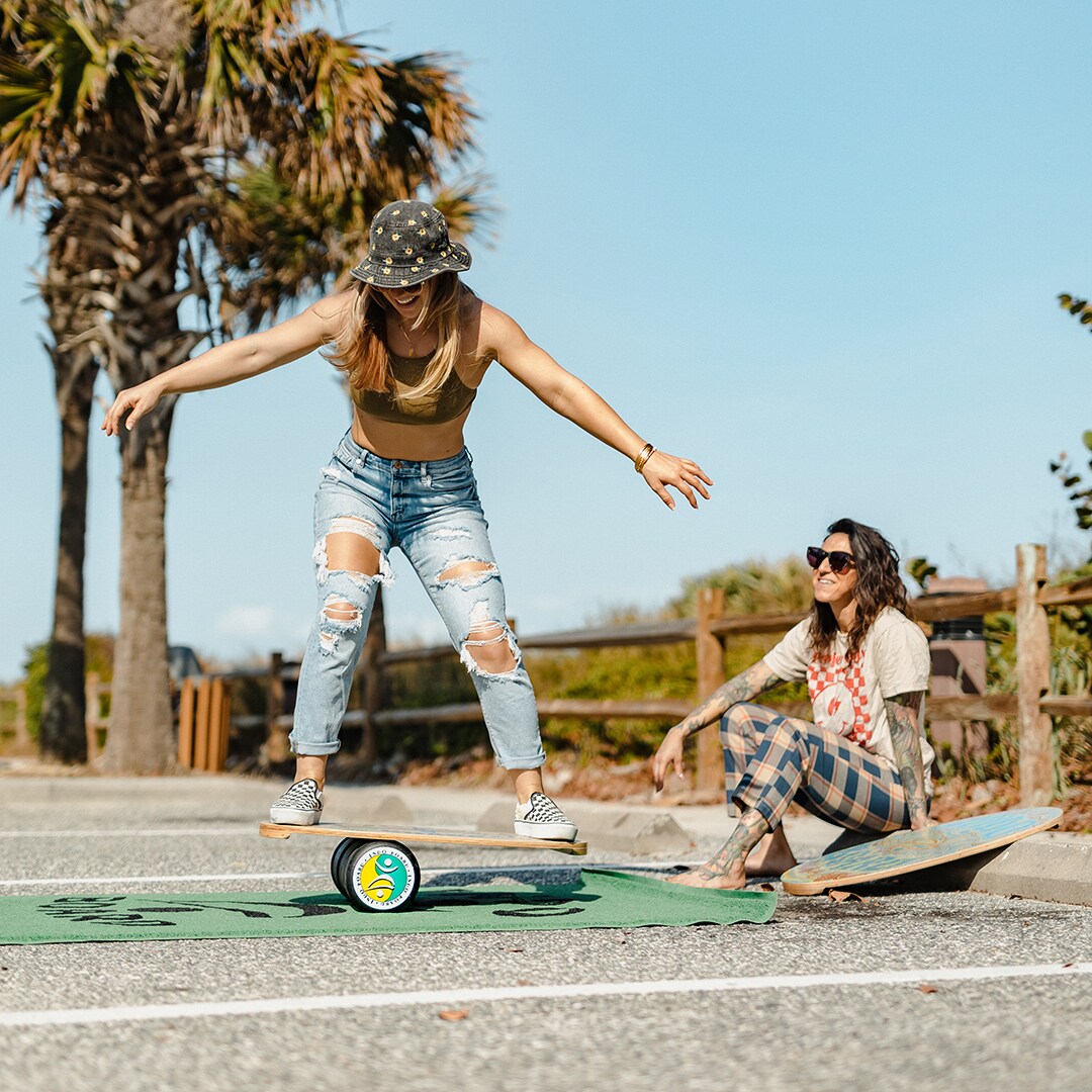 Två kvinnor tränar balans utomhus, en balanserar på en Indoboard med roller på en matta medan den andra tittar på