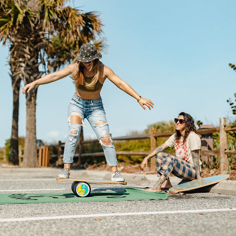 Två kvinnor tränar balans utomhus, en balanserar på en Indoboard med roller på en matta medan den andra tittar på