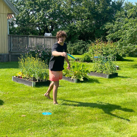 En man kastar en discgolf-frisbee barfota på en gräsmatta med pallkragar och trädgårdsland i bakgrunden. Fokus på kaströrelse