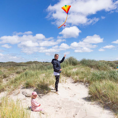 Vuxen och barn som flyger en färgglad Eddy Rainbow drake med lång gul svans och vitt snöre bland sanddyner vid stranden
