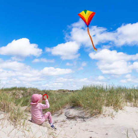 Barn i rosa kläder som sitter i sanddynerna och flyger en Eddy Rainbow drake med långa orangea svansar och vitt snöre mot en klarblå himmel med moln