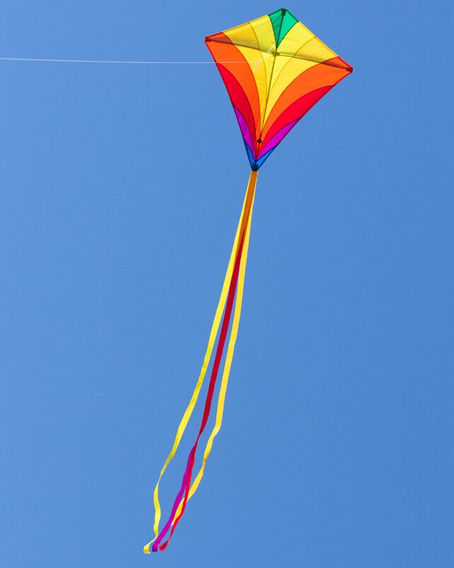 Närbild på den färgglada Eddy Rainbow-draken som flyger mot en klarblå himmel med sina långa gula och röda svansar och vitt snöre