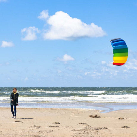 En ung kvinna står på en sandstrand vid havet och flyger en stor regnbågsfärgad Symphony-drake mot en blå himmel med vita moln.