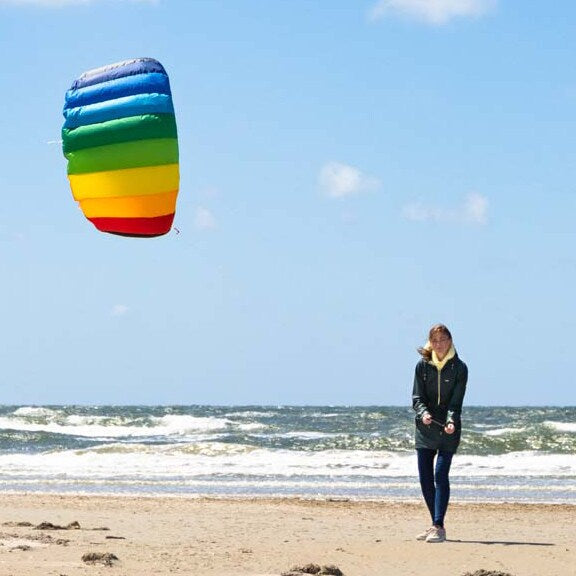Gemini sa En kvinna som flyger en regnbågsfärgad Symphony-drake på en sandstrand vid havet en solig dag.