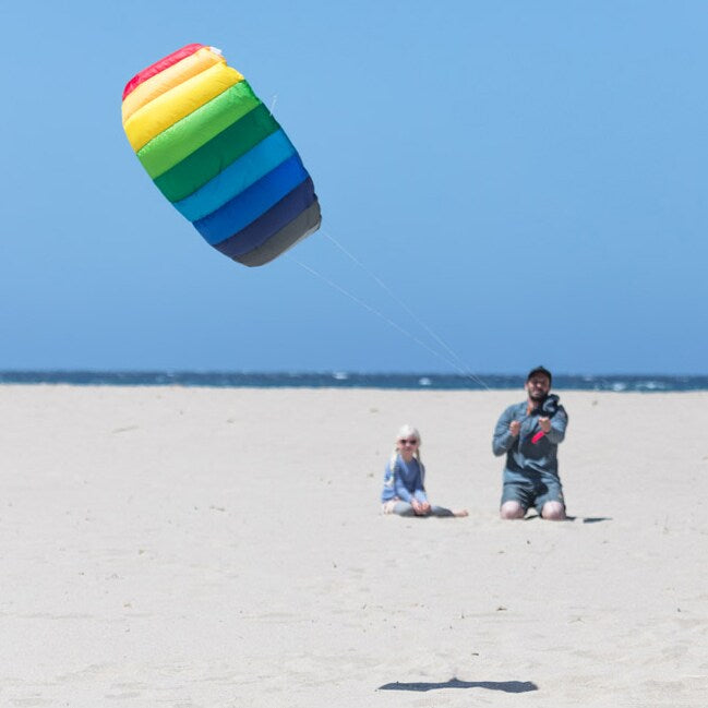 En man och ett barn sitter på en sandstrand och flyger en stor regnbågsfärgad Symphony-drake mot en klarblå himmel.