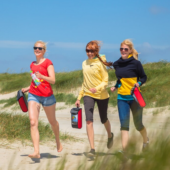 Tre personer som springer skrattande längs en sandstrand bärandes på varsin röd väska innehållande en Symphony Beach III-drake