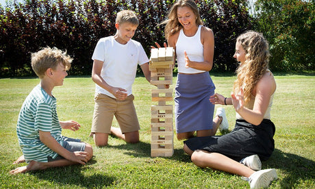 Fyra personer spelar Giant Tower (Stort tornspel) från BEX utomhus på en gräsmatta. En kvinna ska dra ut en kloss ur det höga trätornet