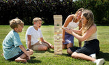 Två pojkar och två flickor spelar Giant Tower från BEX utomhus på en gräsmatta. En av pojkarna gapar av spänning medan en flicka drar ut en kloss
