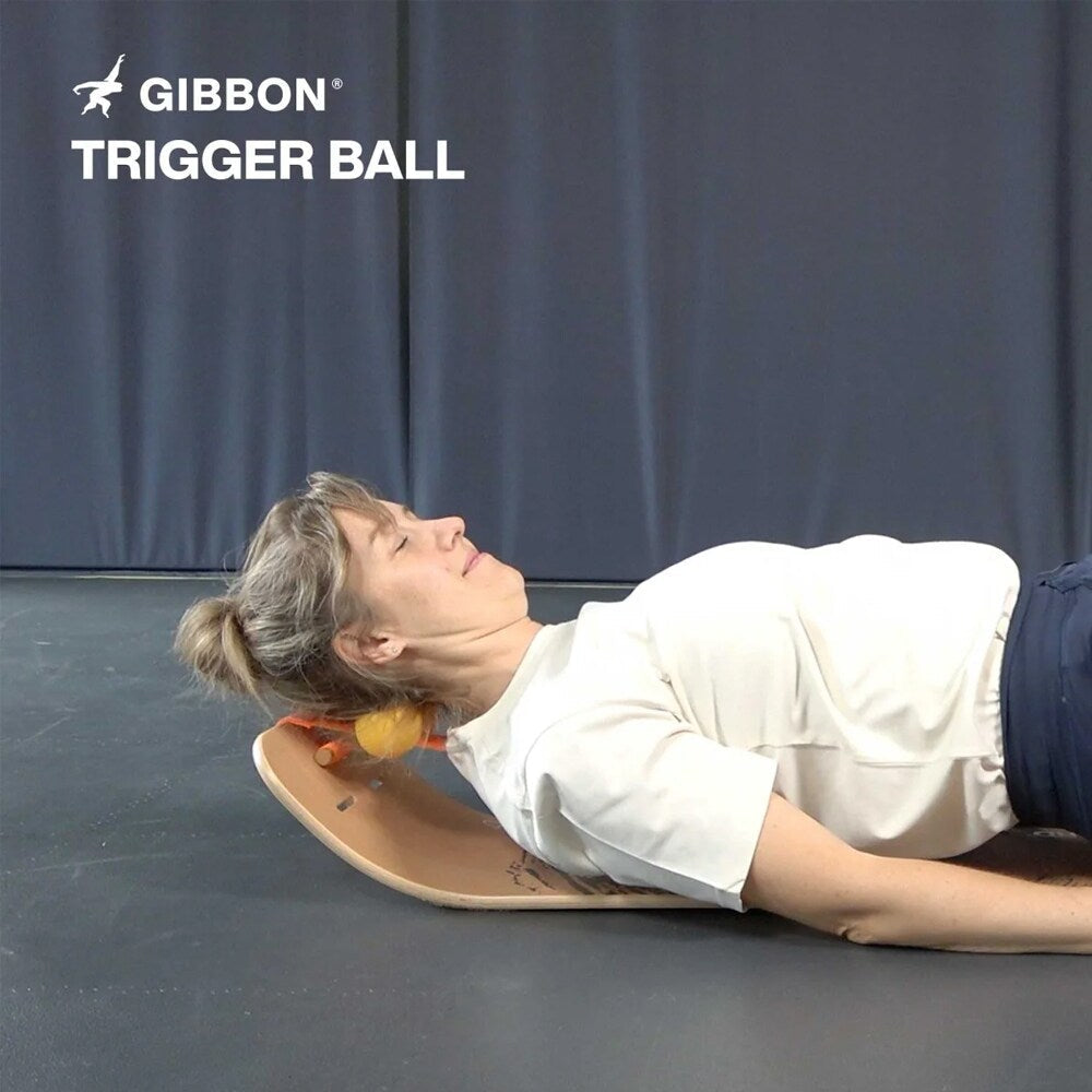 En kvinna som ligger på rygg på en GIBBON balansbräda och vilar nacken mot en gul GIBBON Trigger Ball för avlastning och massage.