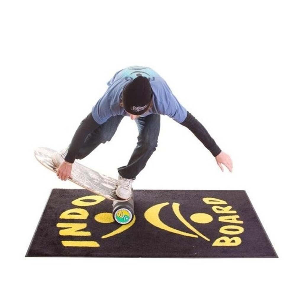 En man balanserar på en Indo Board Rockerboard på en svart Indo Board-matta. Mattan har gul logotyp och brädan ligger på rullen
