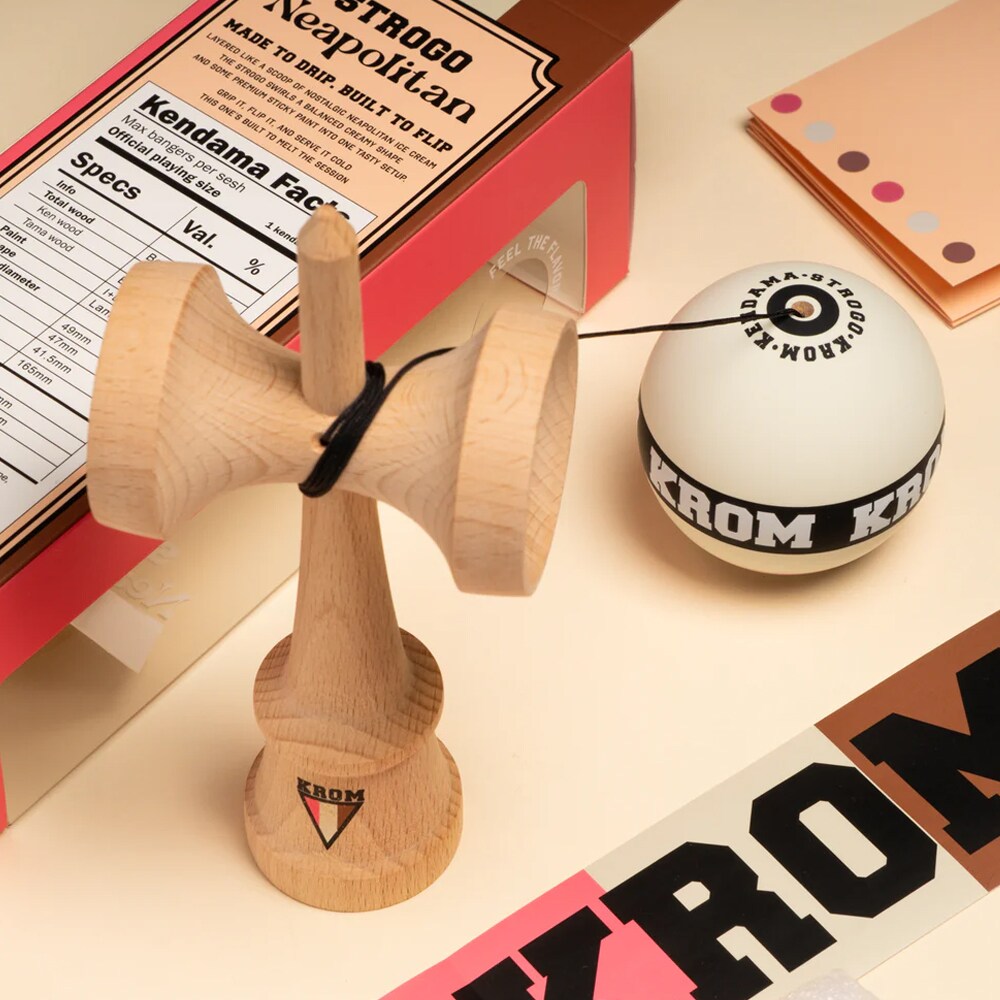 KROM Strogo Neapolitan kendama i ljust trä bredvid sin rosa och brunmönstrade förpackning, omgiven av tillbehör som snöre och klistermärken på en beige yta.