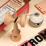 KROM Strogo Neapolitan kendama i ljust trä bredvid sin rosa och brunmönstrade förpackning, omgiven av tillbehör som snöre och klistermärken på en beige yta.