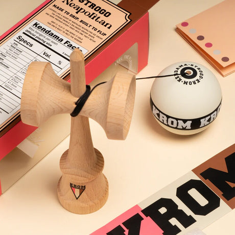 KROM Strogo Neapolitan kendama i ljust trä bredvid sin rosa och brunmönstrade förpackning, omgiven av tillbehör som snöre och klistermärken på en beige yta.