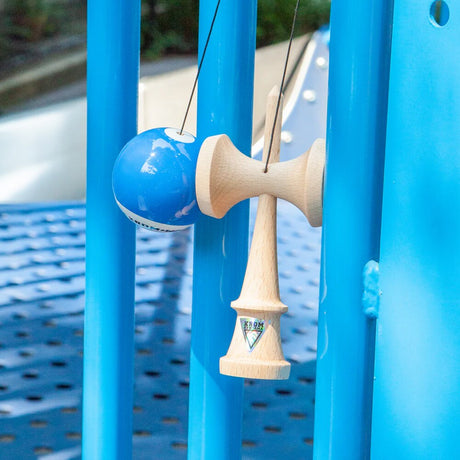 KROM Pop LOL kendama i ljust bokträ med en blå boll (tama) och vitt snöre hängande från ett blått klätterställnng utomhus