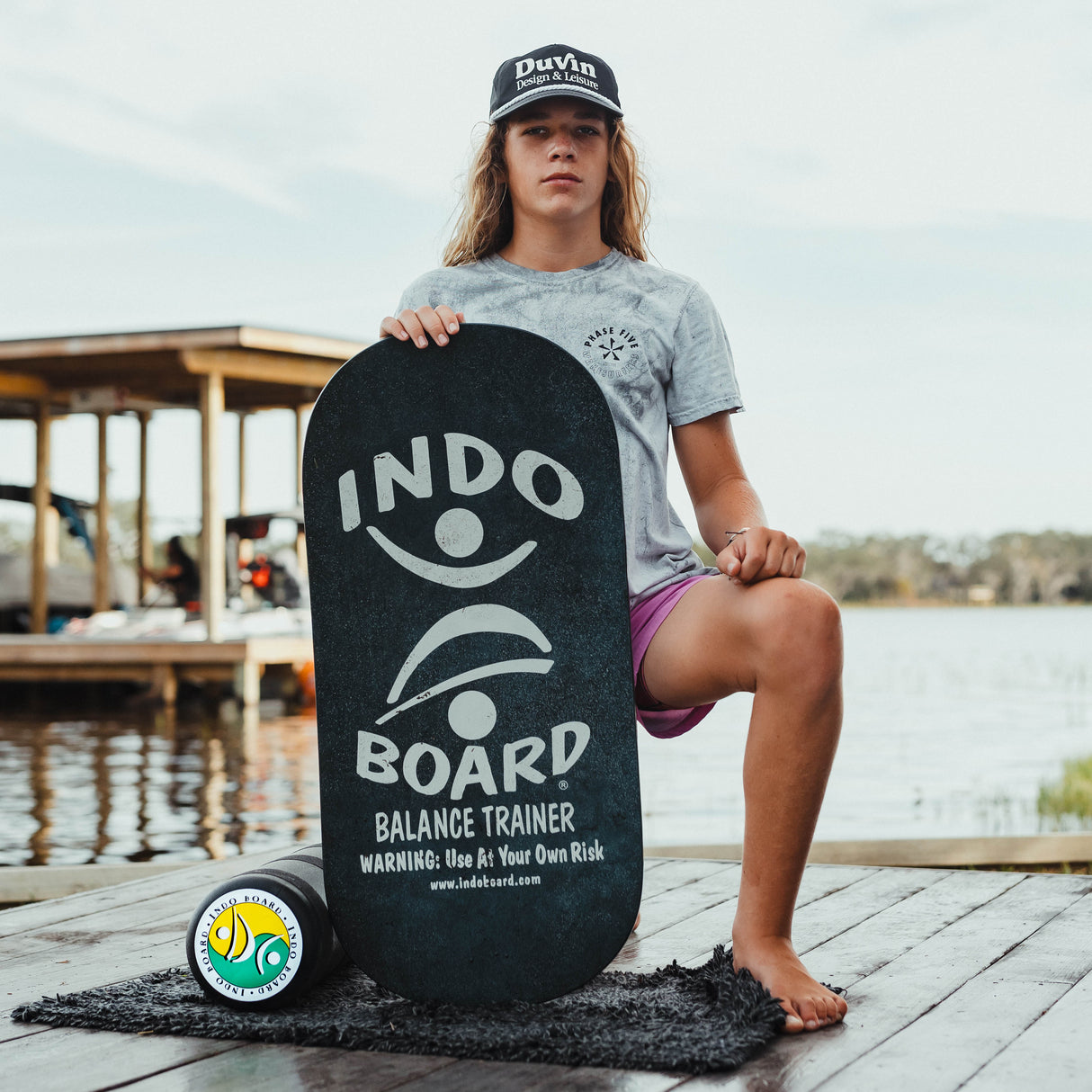 Indo Board Rockerboard i svart med vit logotyp hålls av en person som sitter på en brygga. Visar den svarta rullen och en liten matta