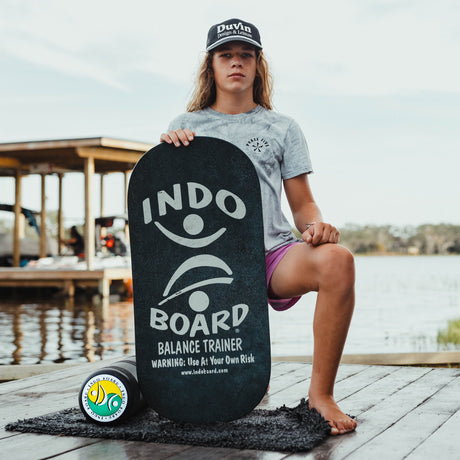 Indo Board Rockerboard i svart med vit logotyp hålls av en person som sitter på en brygga. Visar den svarta rullen och en liten matta