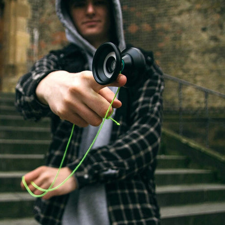 Närbild på en person i grå hoodie och rutig skjorta som balanserar en svart jojo på fingret med ett neon-grönt snöre. Personen står utomhus framför en stentrappa.