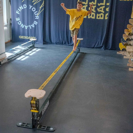 En pojke som balanserar barfota på en GIBBON Slackrack med gul slackline i en träningslokal, där sittytor i trä är monterade i båda ändar av stålramen.