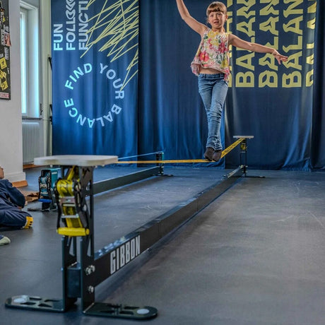 En ung flicka balanserar på en svart GIBBON Slackrack med gul slackline i en träningslokal, där en sittyta i trä är monterad på stålramens främre stolpe.