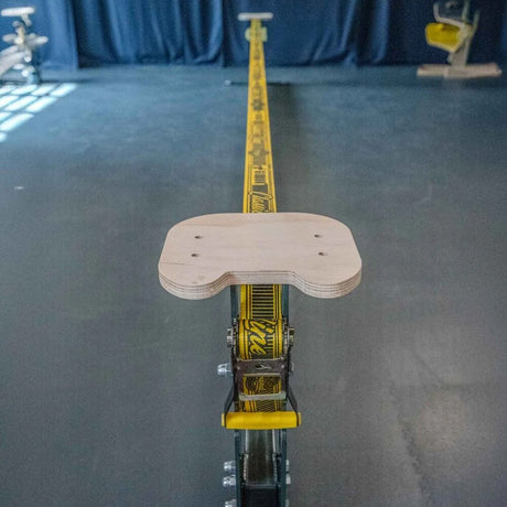 En GIBBON Slackrack sedd uppifrån längs den gula slacklinen, med fokus på en monterad sittyta i ljust trä ovanpå spärrhaken.