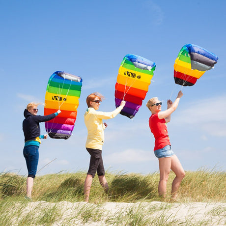 Tre kvinnor på en sanddyn som håller i varsin färgglad Symphony Beach paragliderdrake med vitt snöre mot en klarblå himmel