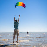 Man som hoppar i vattenbrynet på en strand och styr en färgglad Symphony Beach paragliderdrake med vitt snöre mot en blå himmel