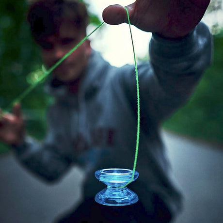 En person i en grå hoodie som gör ett jojo-trick utomhus med en genomskinlig blå Arrow jojo. Jojon hänger längst ner i ett neongrönt snöre medan personen håller i den andra änden
