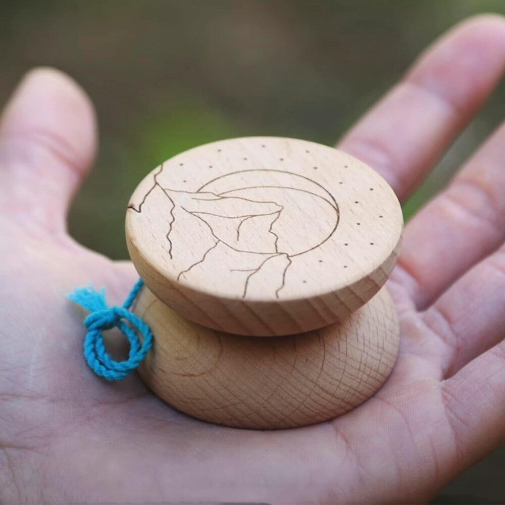 Närbild på en Boho Wooden jojo i ljust trä som ligger i en handflata. Jojons sida har en ingraverad design föreställande berg, en måne och små prickar som stjärnor. Ett blått snöre är knutet vid jojon.