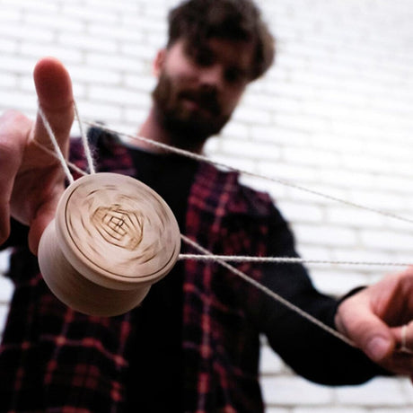 Närbild på en person som utför ett jojo-trick med en roterande Boho Wooden jojo i trä. Personen bär en rutig skjorta och balanserar jojon i ett vitt snöre framför en vit tegelvägg