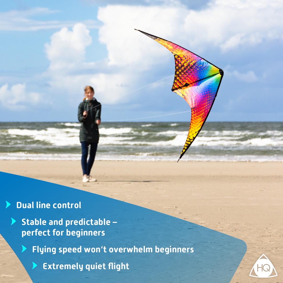 Färgstark styrbar Bebop-drake med ett prickigt regnbågsmönster mot en svart bas som flyger i luften över en sandstrand med en kvinna i bakgrunden