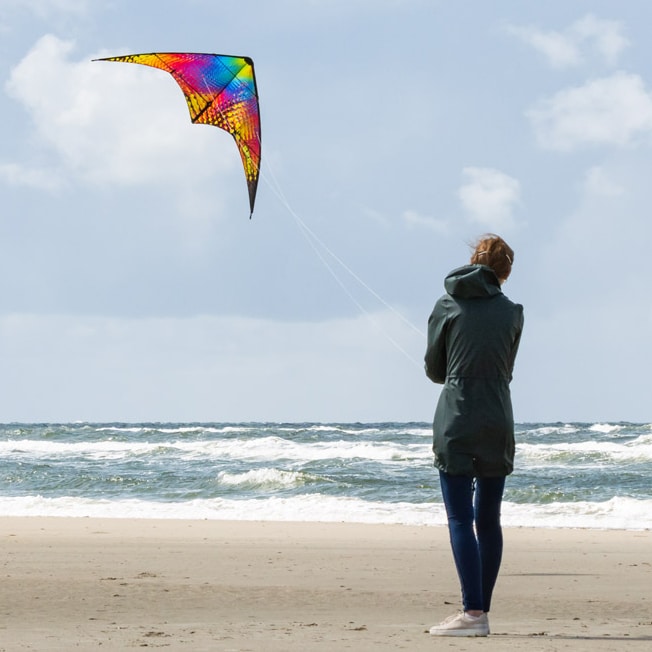 Färgstark styrbar Bebop-drake med ett mönster av gula, orangea, lila och gröna cirklar mot en svart bas som flyger i luften vid kusten med en kvinna i bakgrunden