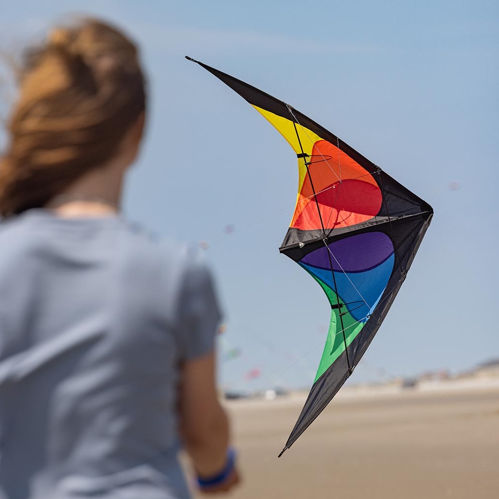 Färgstark styrbar Bebop-drake med ett mönster av röda, gula, lila och gröna cirklar mot en svart bas som flyger i luften över en sandstrand med en person i bakgrunden