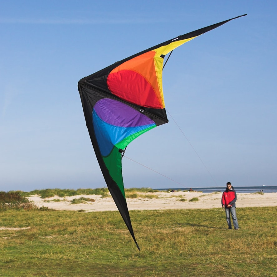 En stor styrbar drake av märket Bebop med ett färgstarkt segel i rött, orange, lila, blått och grönt mot en svart bas som flyger i luften över ett gräsbevuxet dynområde med en person i bakgrunden