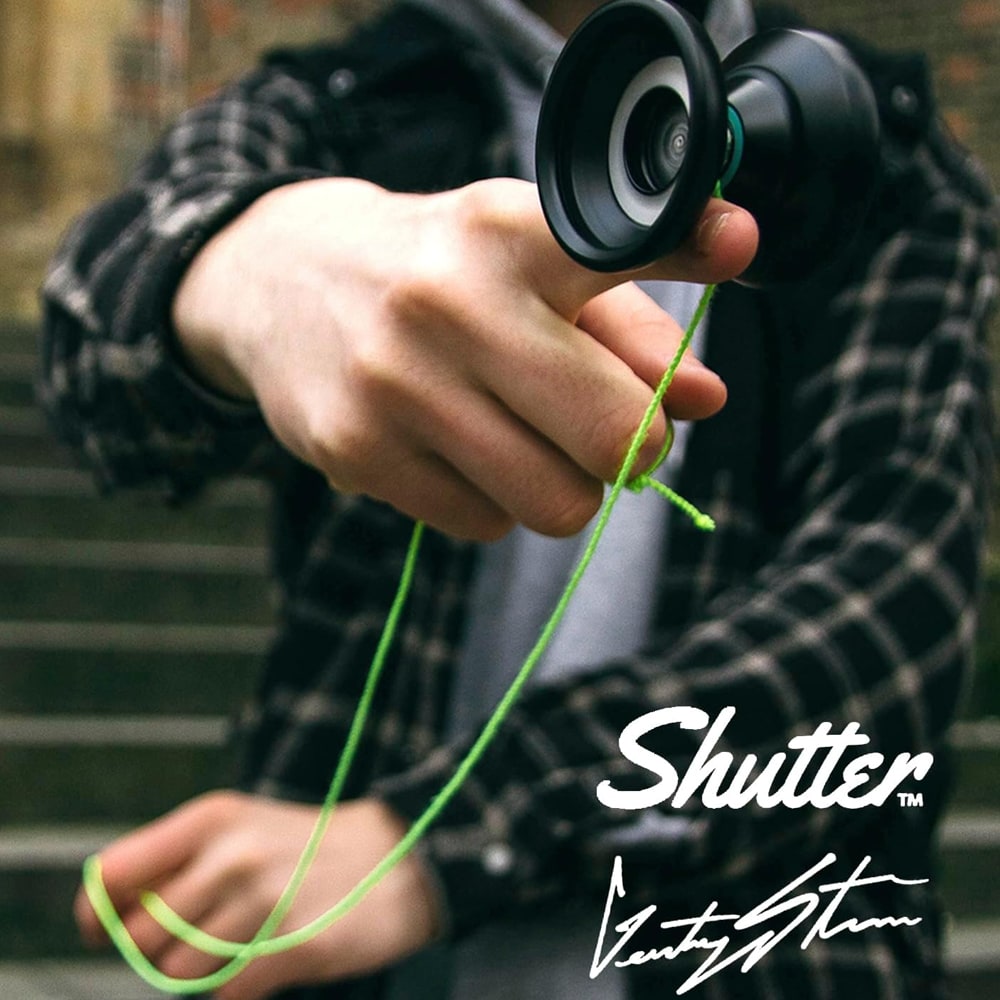 En person i grå hoodie och rutig skjorta håller fram en svart jojo mot kameran i en utomhusmiljö med en stentrappa i bakgrunden. Ett neongrönt snöre löper från jojon ner mot personens andra hand.