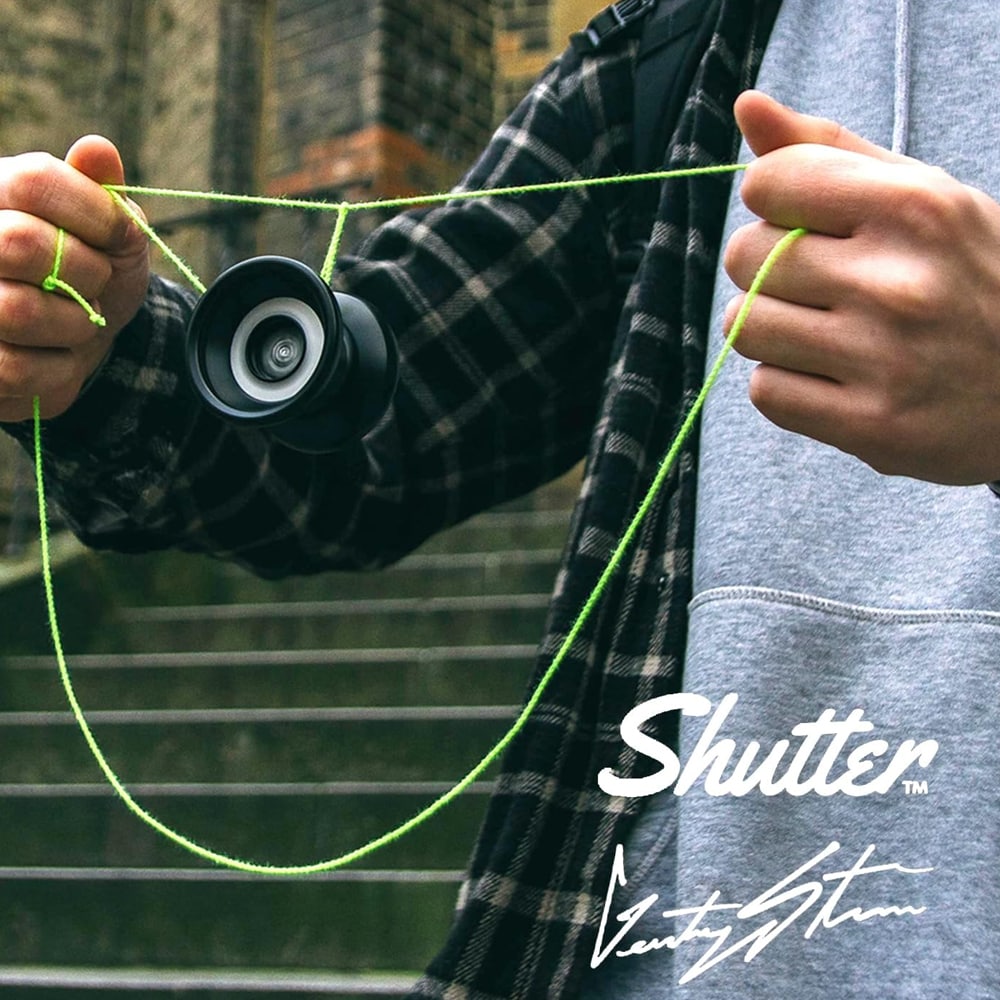 En person i grå hoodie och rutig skjorta sträcker fram en svart jojo mot kameran. Jojon balanserar på personens finger och har ett neongrönt snöre som hänger ner i en ögla. Bakgrunden visar en suddig stentrappa utomhus.