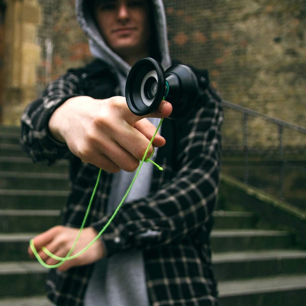 Närbild på en person i grå hoodie och rutig skjorta som balanserar en svart jojo på fingret med ett neon-grönt snöre. Personen står utomhus framför en stentrappa.