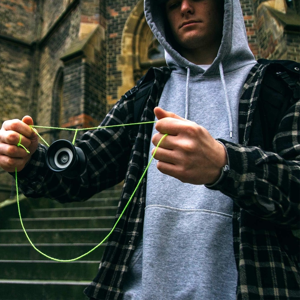 En kille i en grå huvtröja och rutig skjorta som gör ett trick med en svart jojo och ett neongrönt snöre framför en stentrappa och en gammal byggnad