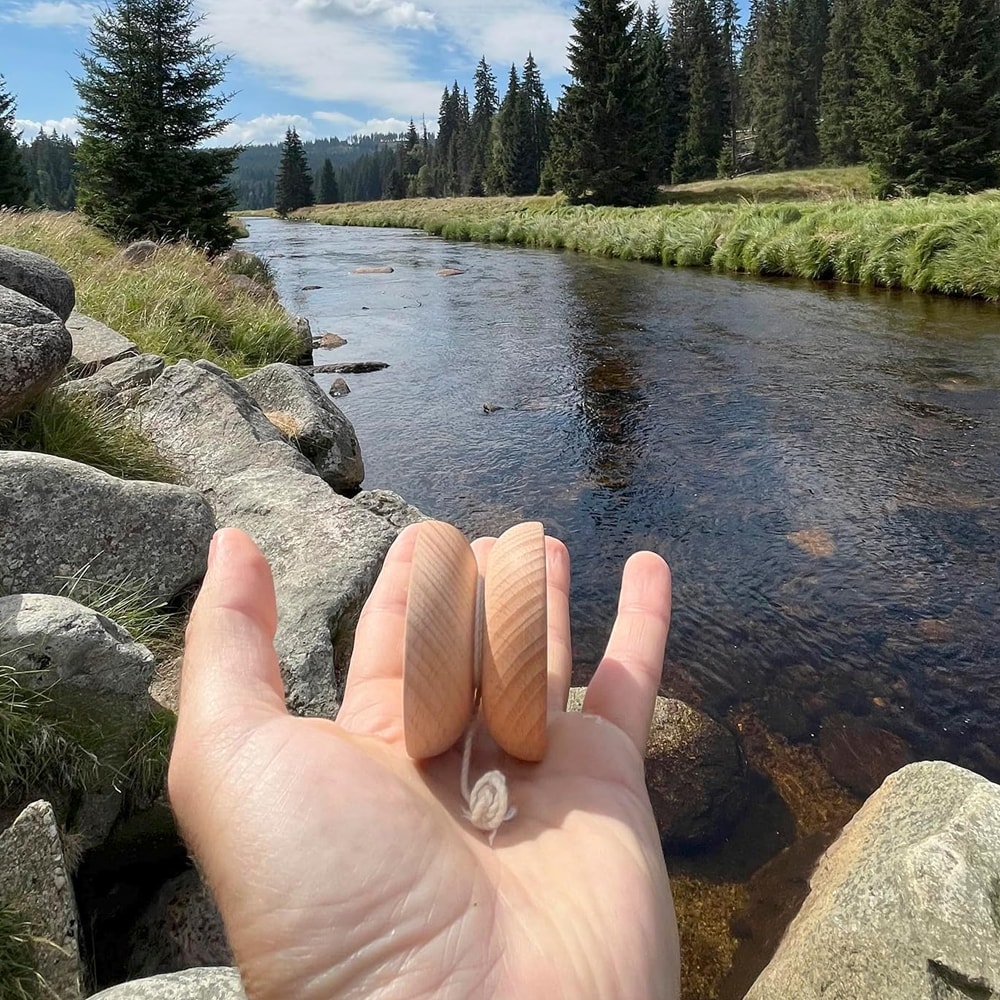 Närbild på en person som håller en Boho Wooden jojo i trä i handflatan framför en naturskön miljö med en rinnande å, stenar och granskog under en blå himmel